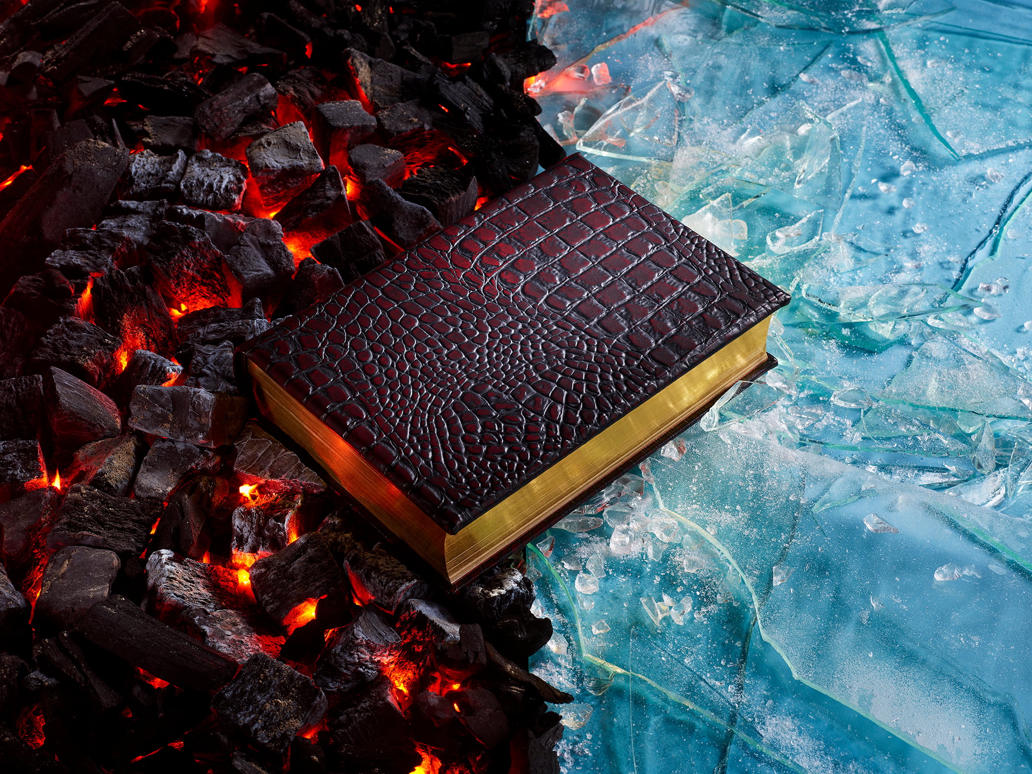 A closed, gold-edged book with a dark textured cover lies between glowing coals on one side and broken ice on the other.