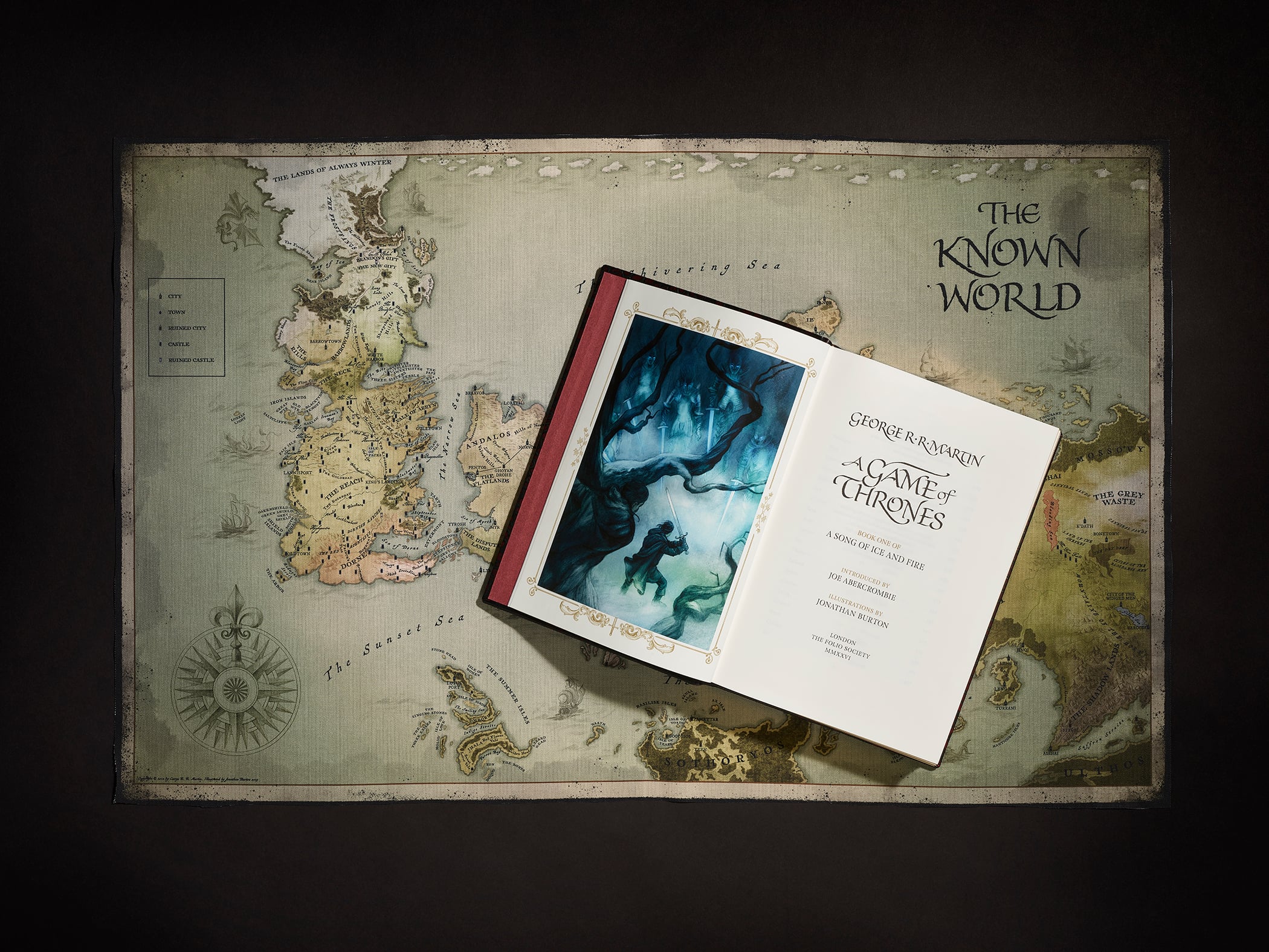 An open "A Game of Thrones" book rests on a map labeled "The Known World," showing an illustrated scene on the left page and the title page on the right.