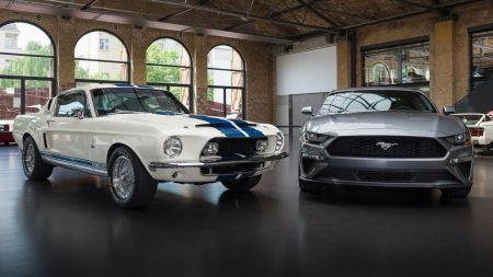 A vintage white Ford Mustang with blue stripes is parked next to a modern silver Ford Mustang inside a building with large arched windows.