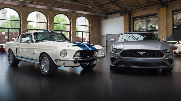 A vintage white Ford Mustang with blue stripes is parked next to a modern silver Ford Mustang inside a building with large arched windows.