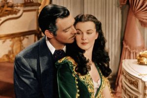 A man in a suit leans in close to a woman in a green dress with gold details as they sit together in an ornate, vintage room with pink and gold decor.