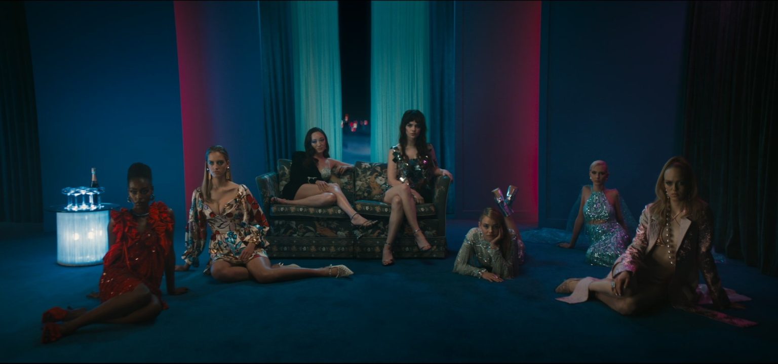 Six women in glamorous, glittery outfits pose on and around a sofa in a dimly lit, stylish room with blue and red lighting.