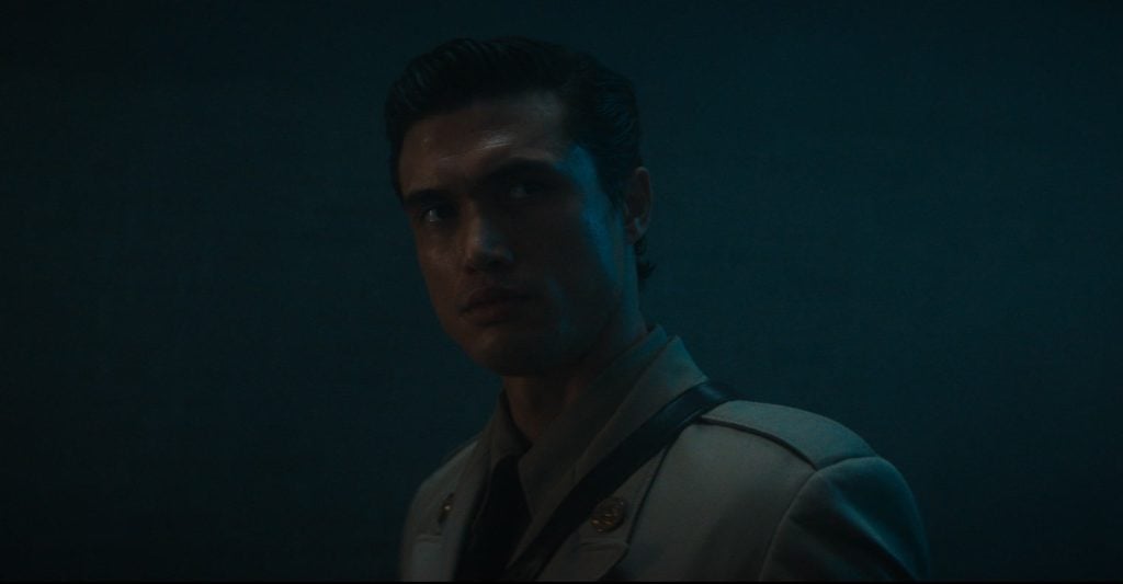 A man in a light-colored military-style uniform stands in a dimly lit, blue-toned room, looking to the side with a serious expression.