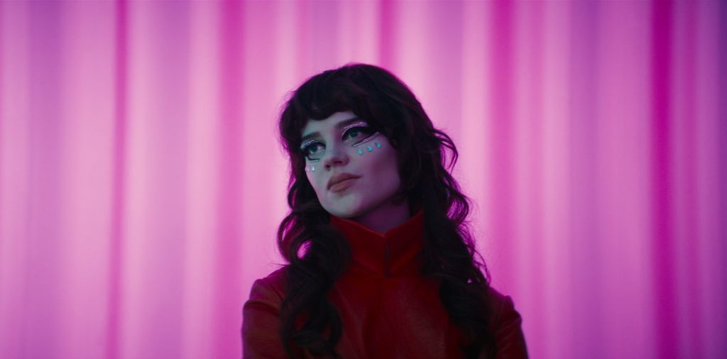 A person with long dark hair and bold eye makeup stands in front of a pink curtain, wearing a red outfit and looking to the side.