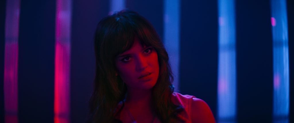 Woman with long brown hair stands under pink and blue lighting, looking slightly to the side with a neutral expression. Vertical light beams are visible in the background.