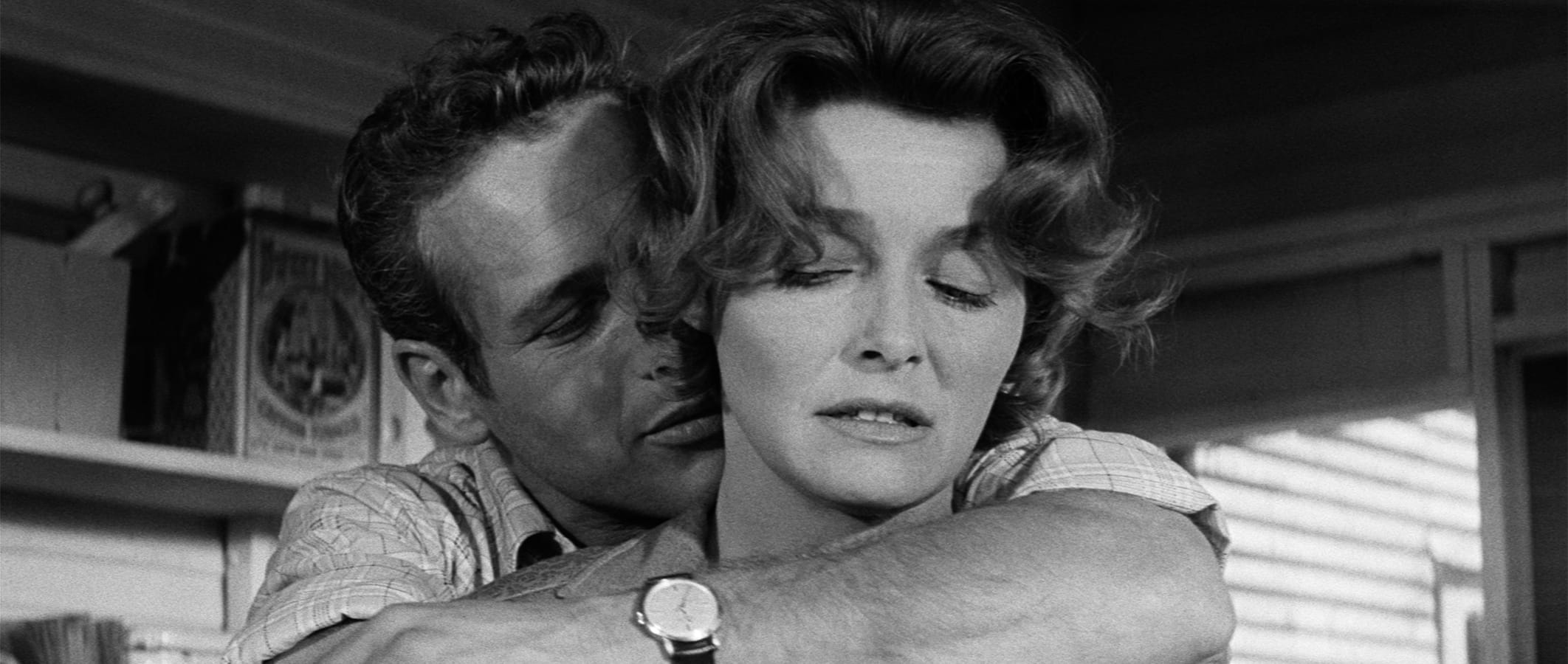 A man embraces a woman from behind indoors; the woman looks away with a neutral expression while the man’s face is close to hers. The image is in black and white.