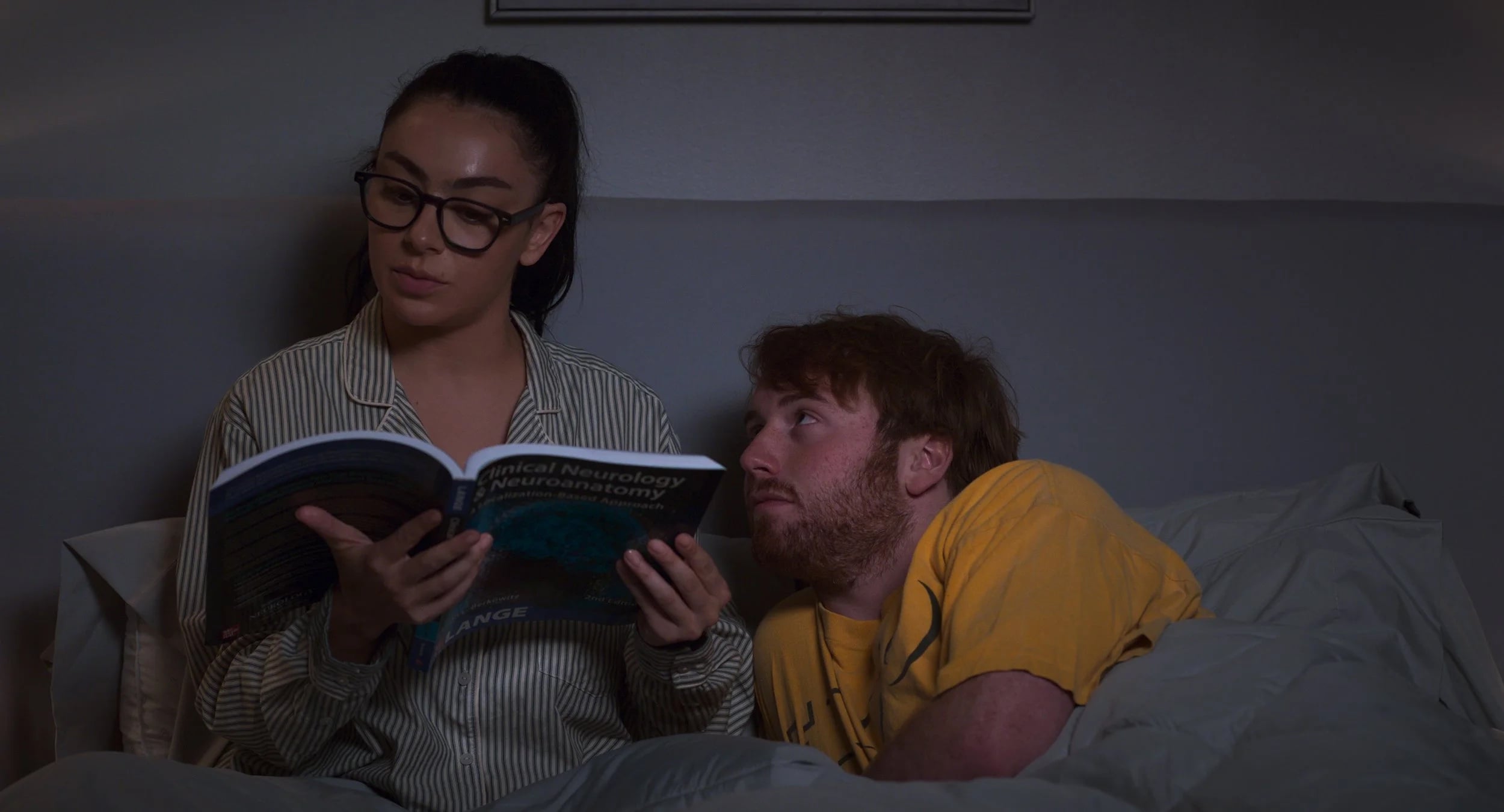 A woman in glasses reads a medical textbook in bed while a man in a yellow shirt lies next to her, looking at her.