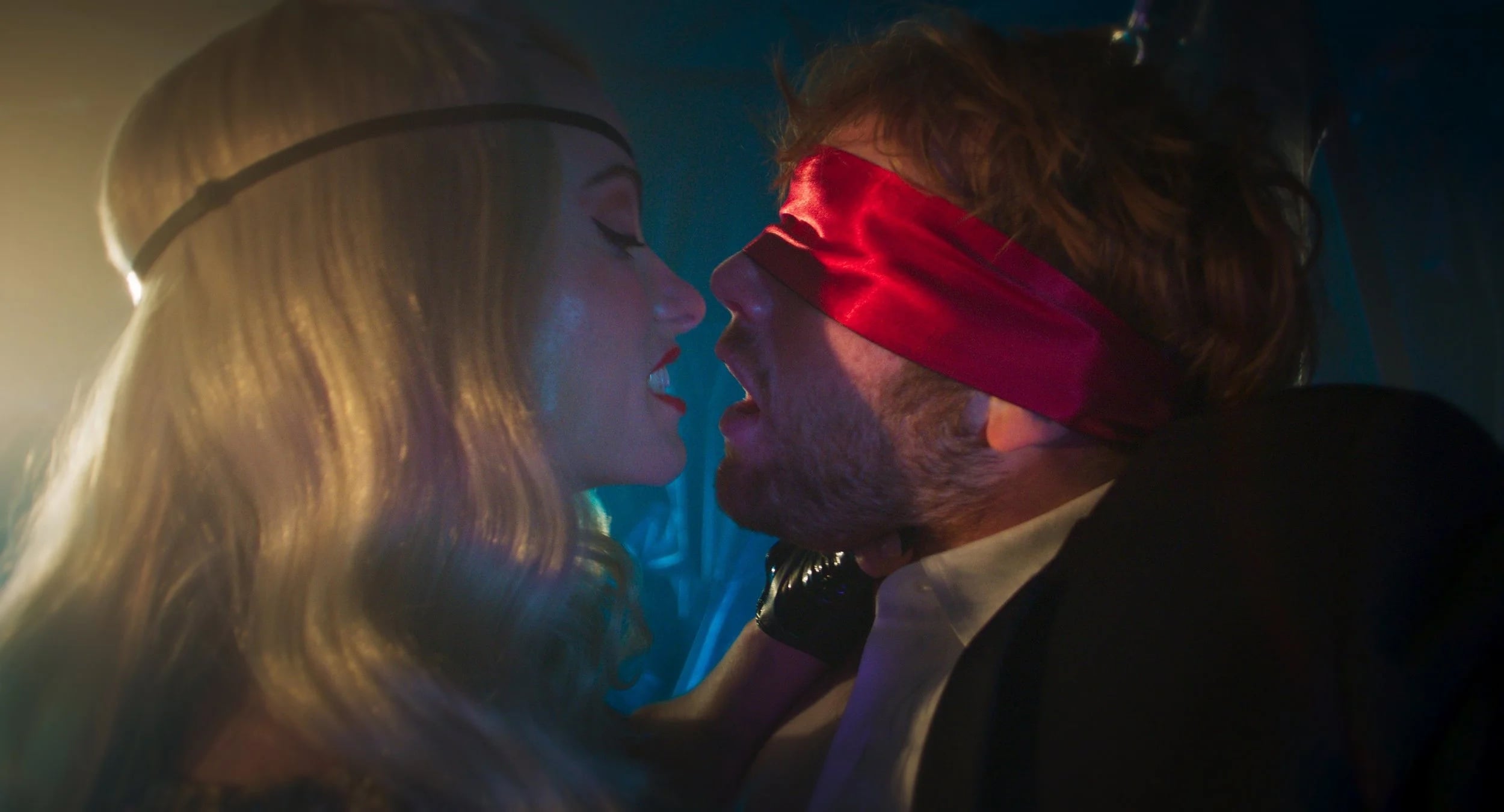 A woman with long blonde hair faces a man in a suit who is blindfolded with a red cloth. They are close together in a dimly lit setting.