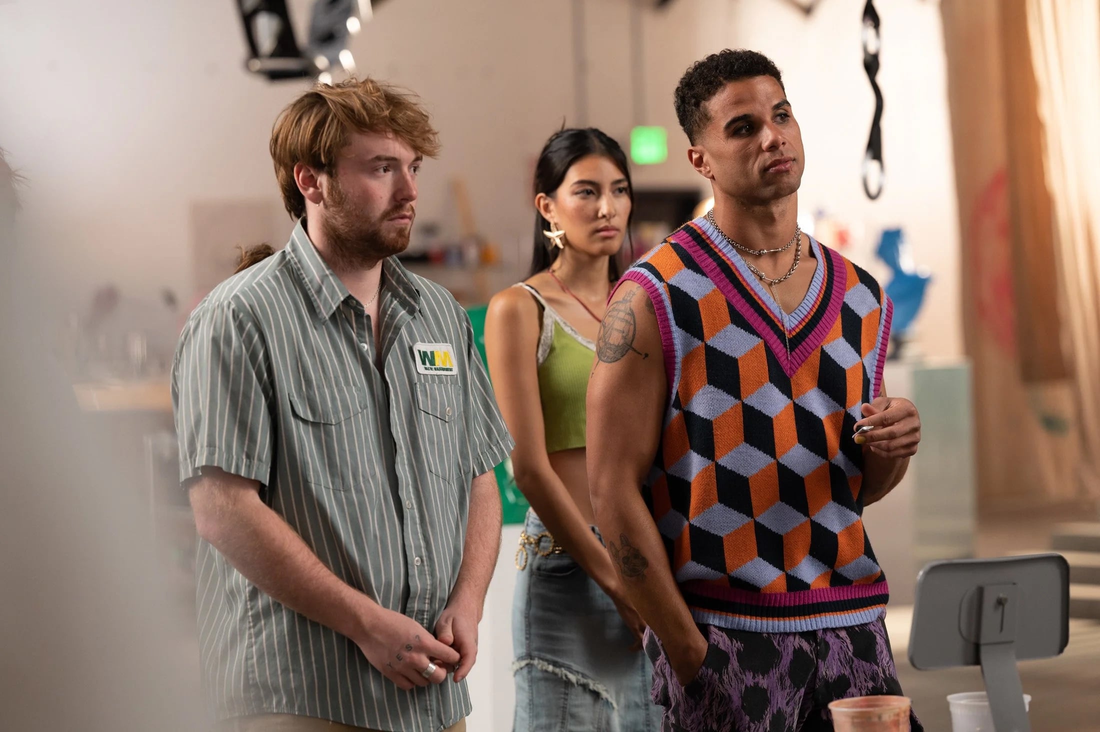 Three people stand indoors, attentively facing forward; one wears a striped shirt, one a green top, and one a colorful geometric vest.