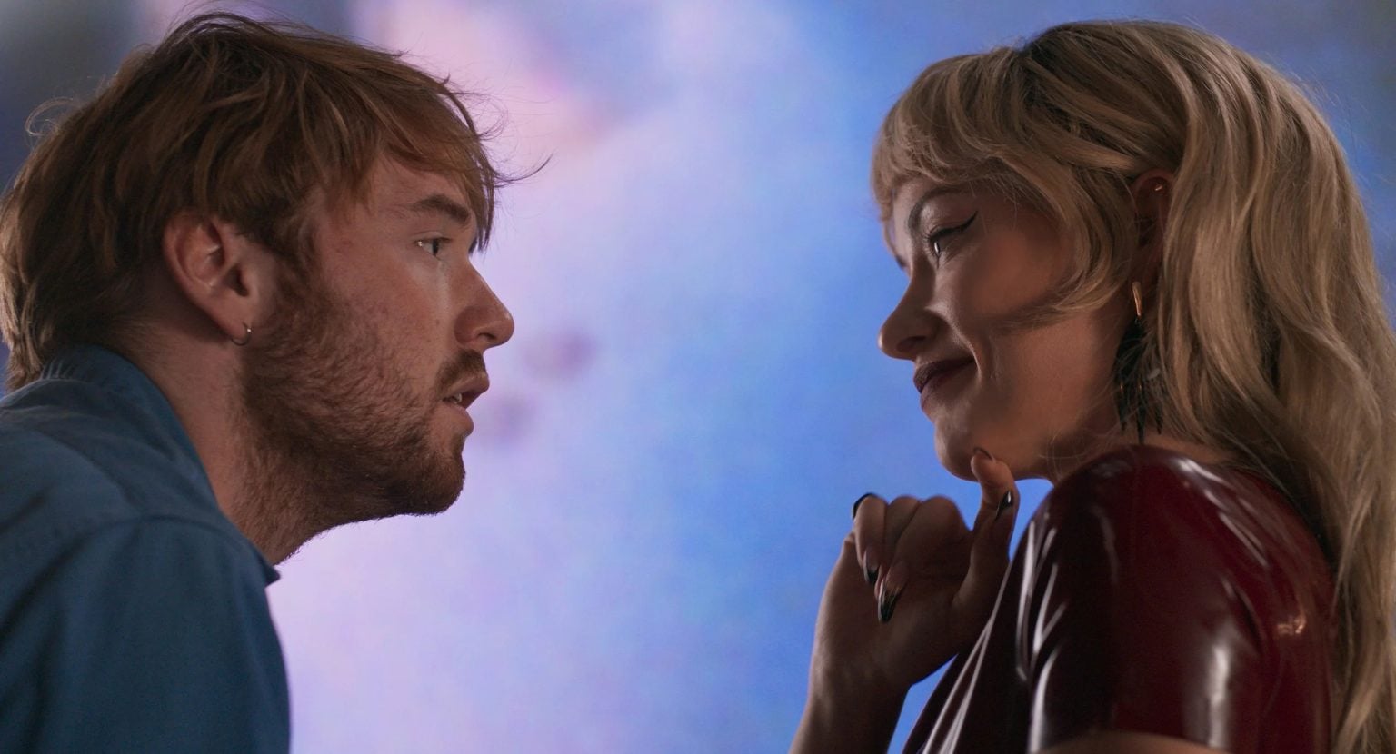 A man with light brown hair faces a woman with blonde hair in a red outfit, both making direct eye contact against a blurred blue and pink background.