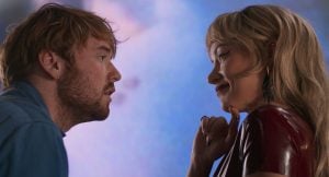 A man with light brown hair faces a woman with blonde hair in a red outfit, both making direct eye contact against a blurred blue and pink background.