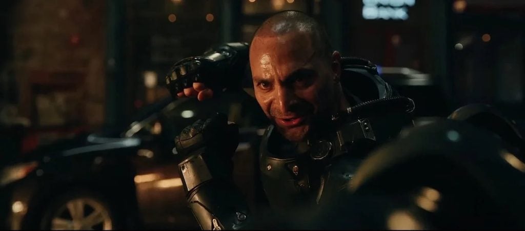 A man in a black armored suit with a shaved head raises his fist, looking intense, with a parked car and blurred lights in the background.