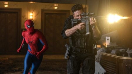 Spider-Man runs behind an armed man firing a rifle inside a dimly lit, damaged room with debris scattered on the floor.