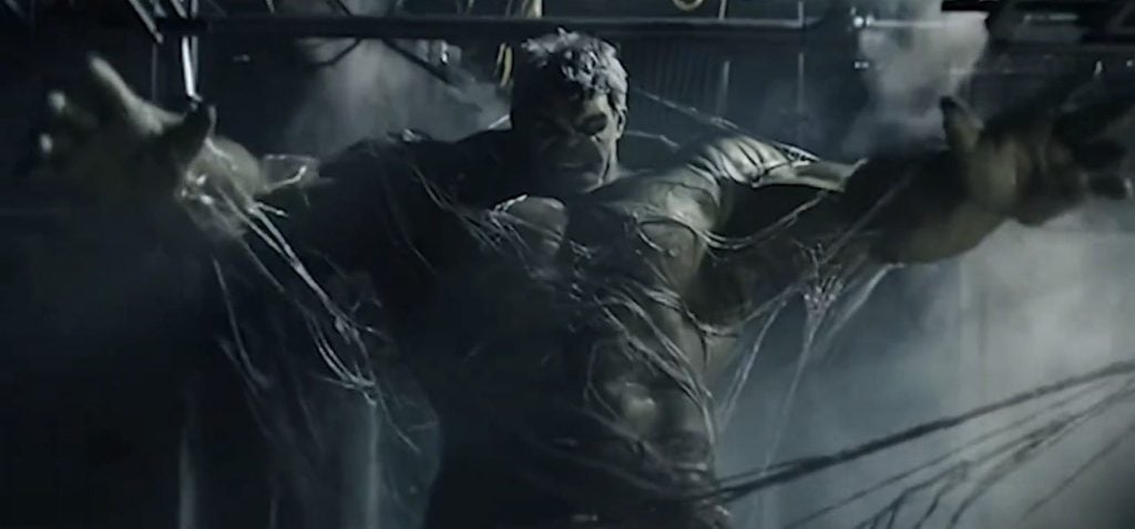 The Hulk stands in a dimly lit room, restrained by multiple thick cables, with smoke and debris in the background.