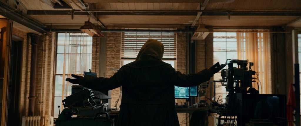A person in a hooded coat stands with arms raised in a cluttered room filled with computer equipment and large windows in the background.