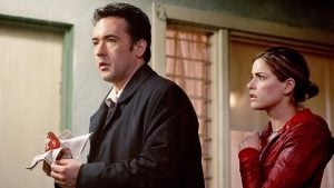 A man holding a paper stands in front of a door, looking concerned, while a woman in a red jacket stands behind him, appearing anxious.