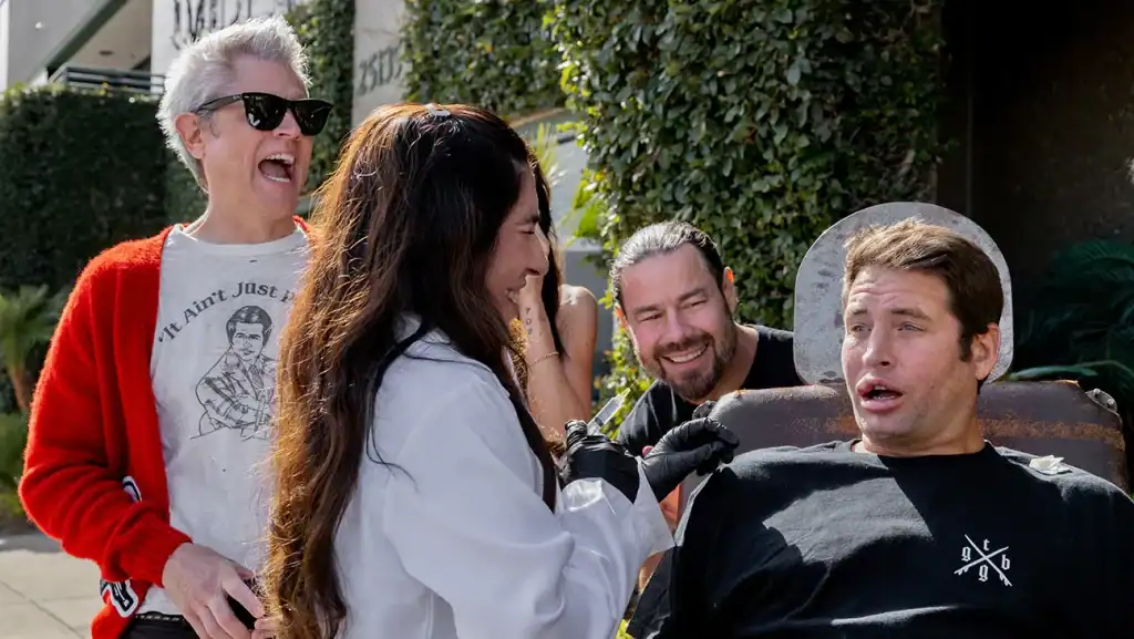 Four people are outdoors; one person sits in a chair looking surprised while another in a lab coat prepares to interact with him, and two others stand nearby laughing.
