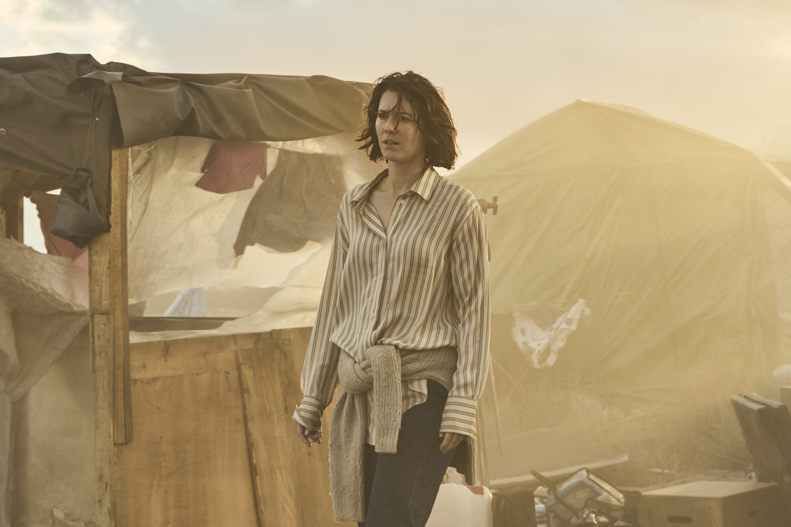 A woman in a striped shirt stands outdoors near makeshift tents and tarps, surrounded by haze and debris.
