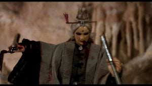 A character in elaborate costume and makeup, with gray hair and decorative hairpieces, holds two weapons and stands against a rocky background.