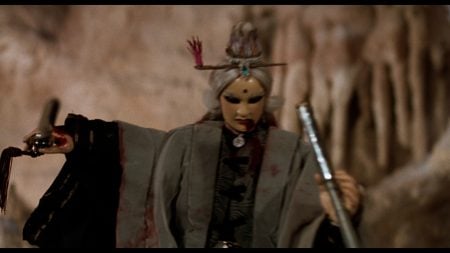 ‘Legend Of The Sacred Stone’ Blu-Ray Review – Martial Arts Mayhem Via Puppets Is A Singular Experience A character in elaborate costume and makeup, with gray hair and decorative hairpieces, holds two weapons and stands against a rocky background.