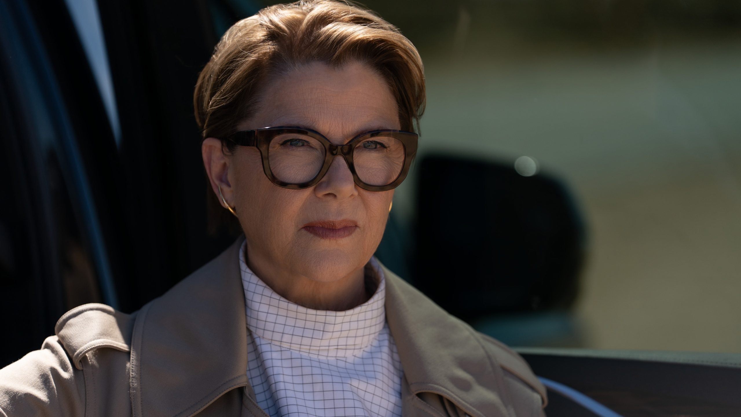 A woman with short hair and glasses sits outdoors, wearing a tan coat and white patterned turtleneck, looking ahead with a serious expression.