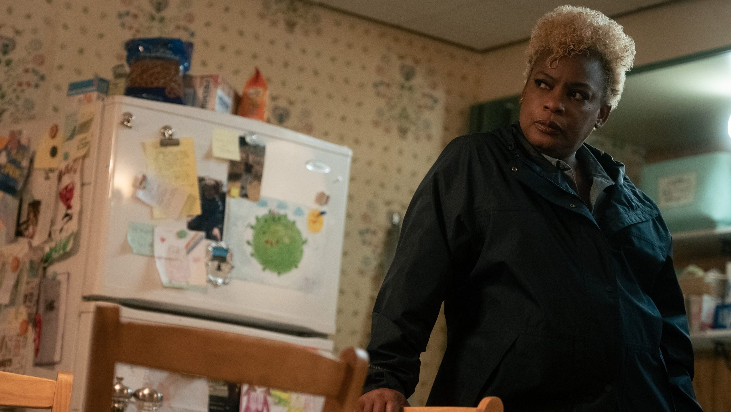 A person in a dark jacket stands in a kitchen, leaning on a table. The fridge behind them is covered in notes and magnets. Shelves and food items are visible in the background.