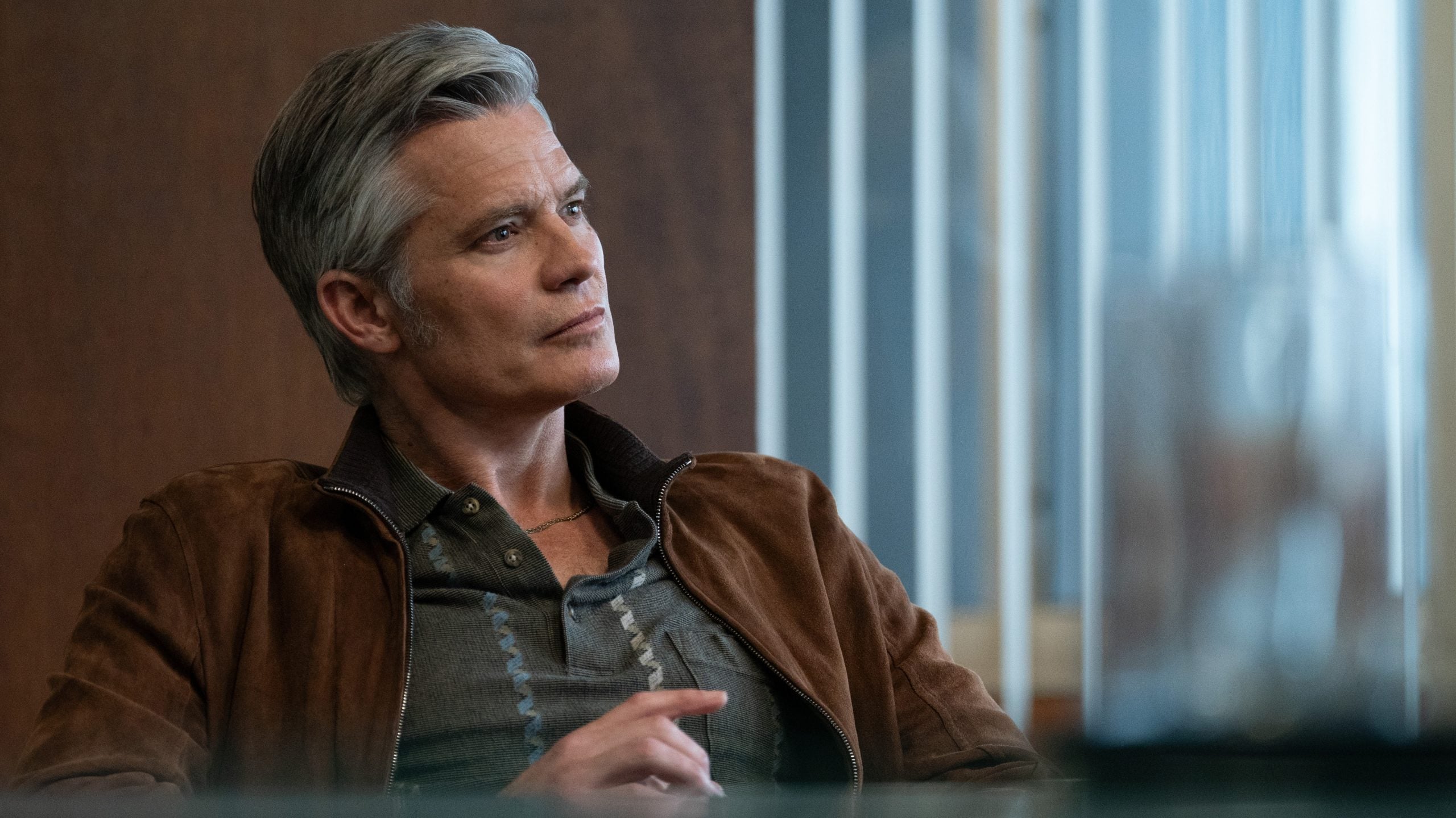A man with gray hair sits indoors, wearing a brown jacket over a dark shirt, looking to the side with a thoughtful expression.