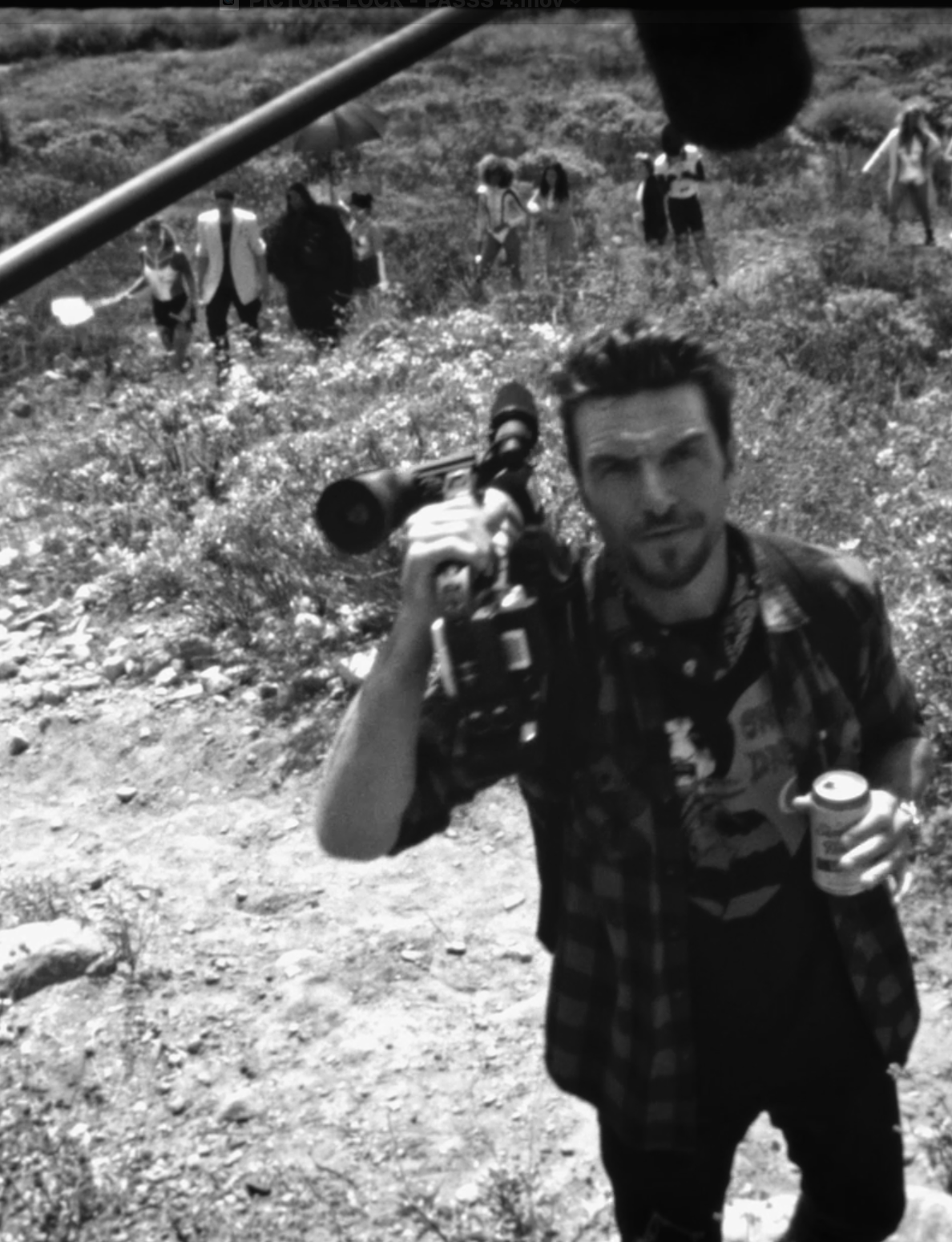 A man holds a camera and a can while looking up, with a boom mic above and a group of people standing in the field behind him.