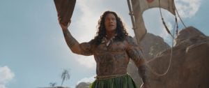 Muscular man with long hair and full-body tattoos holds a wooden oar while standing outdoors with a blue sky and rocks in the background.