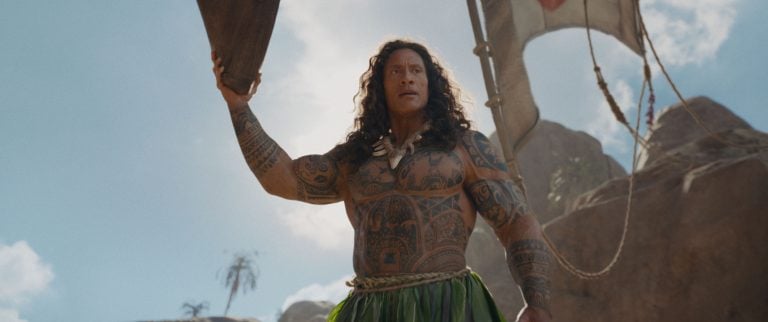 Muscular man with long hair and full-body tattoos holds a wooden oar while standing outdoors with a blue sky and rocks in the background.