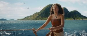 A young woman stands on a boat holding a rope, with the ocean and a lush, mountainous island in the background.