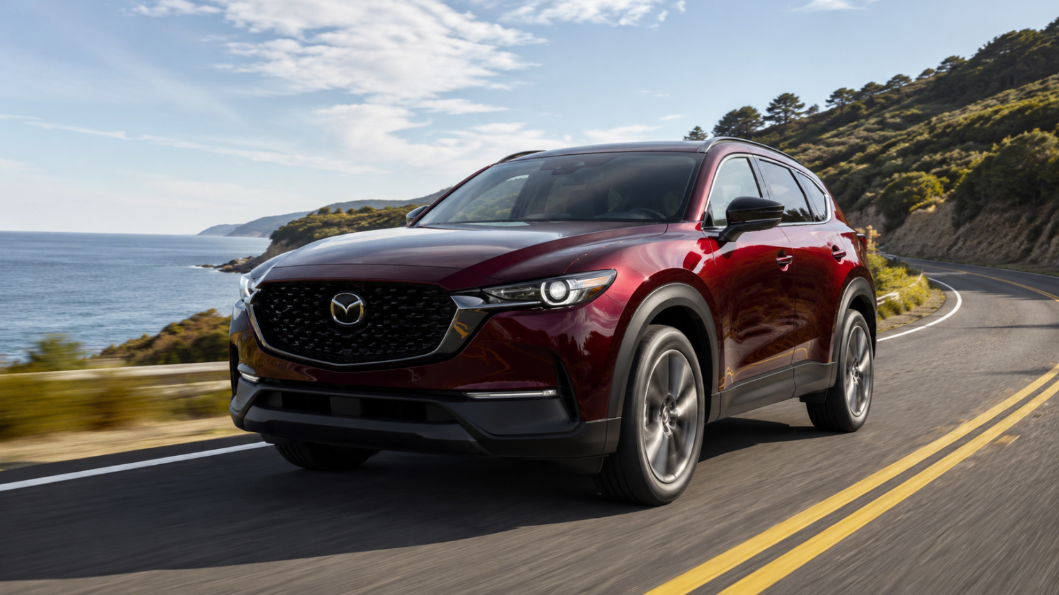 A red Mazda SUV drives along a coastal highway with hills, trees, and the ocean visible in the background under a partly cloudy sky.