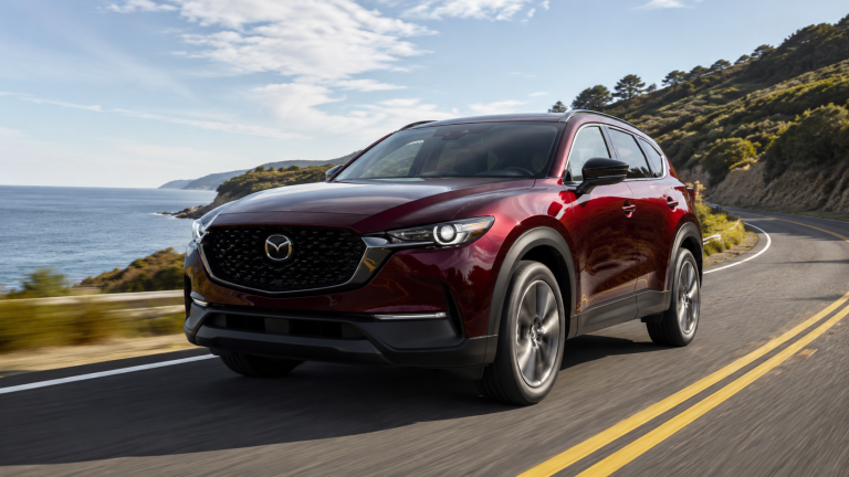 A red Mazda SUV drives along a coastal highway with hills, trees, and the ocean visible in the background under a partly cloudy sky.