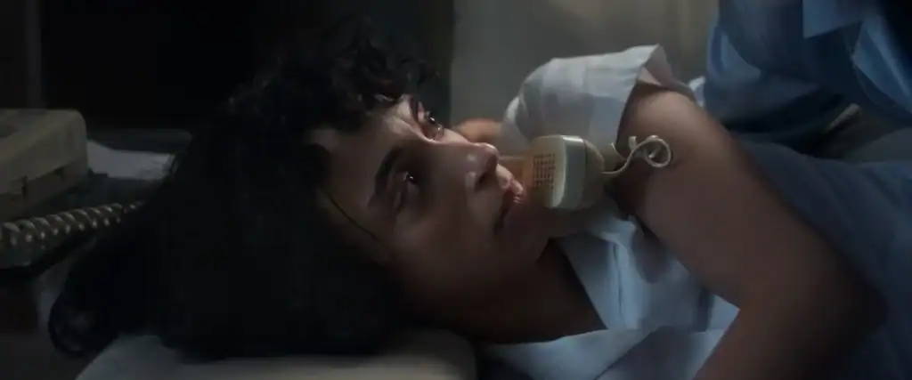 A woman with curly hair lies on her side holding a beige corded telephone to her ear, looking upward with a serious expression.