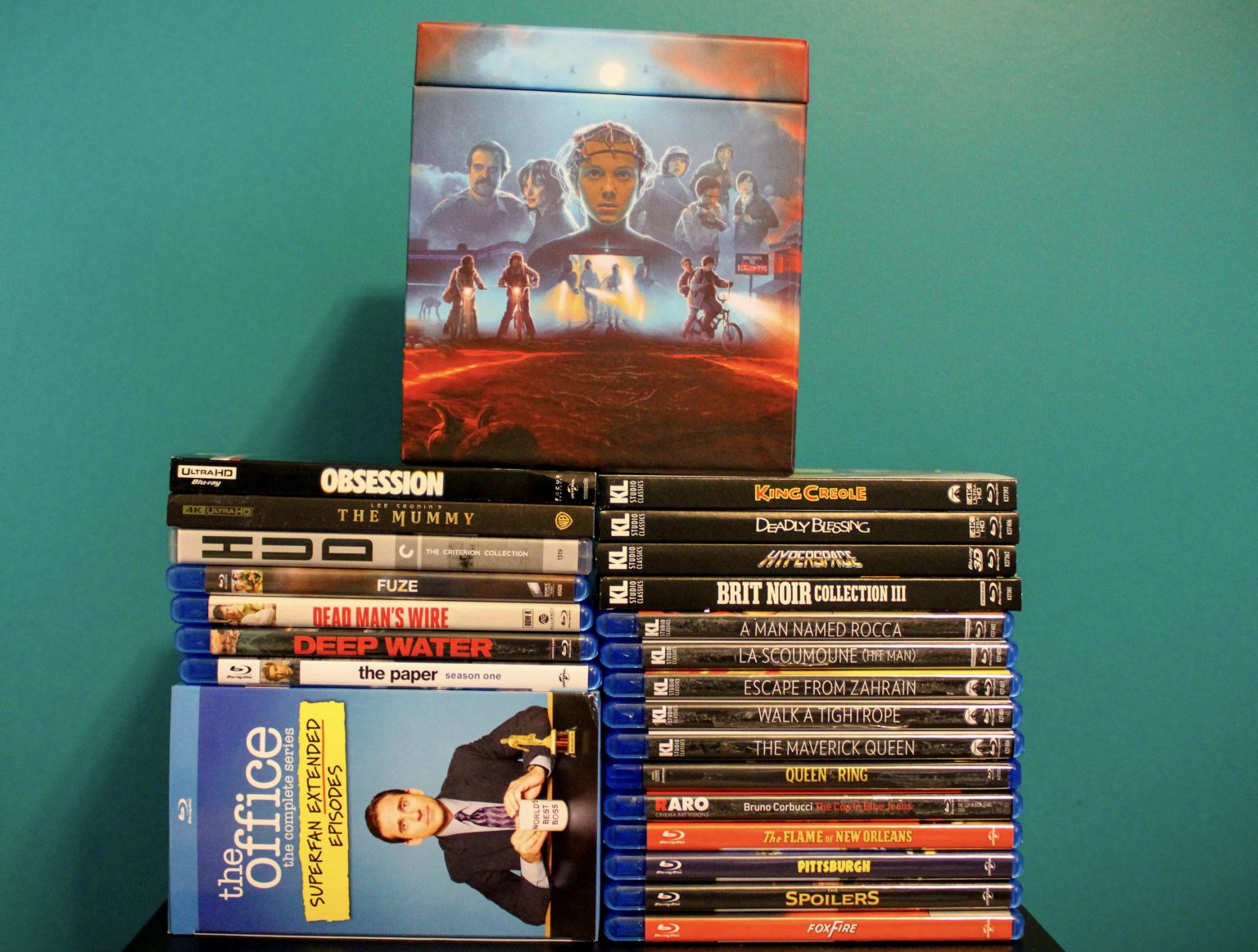A stack of DVD and Blu-ray cases, including "The Office," is arranged in front of a "Dune" collector’s box with illustrated characters, all set against a teal wall.