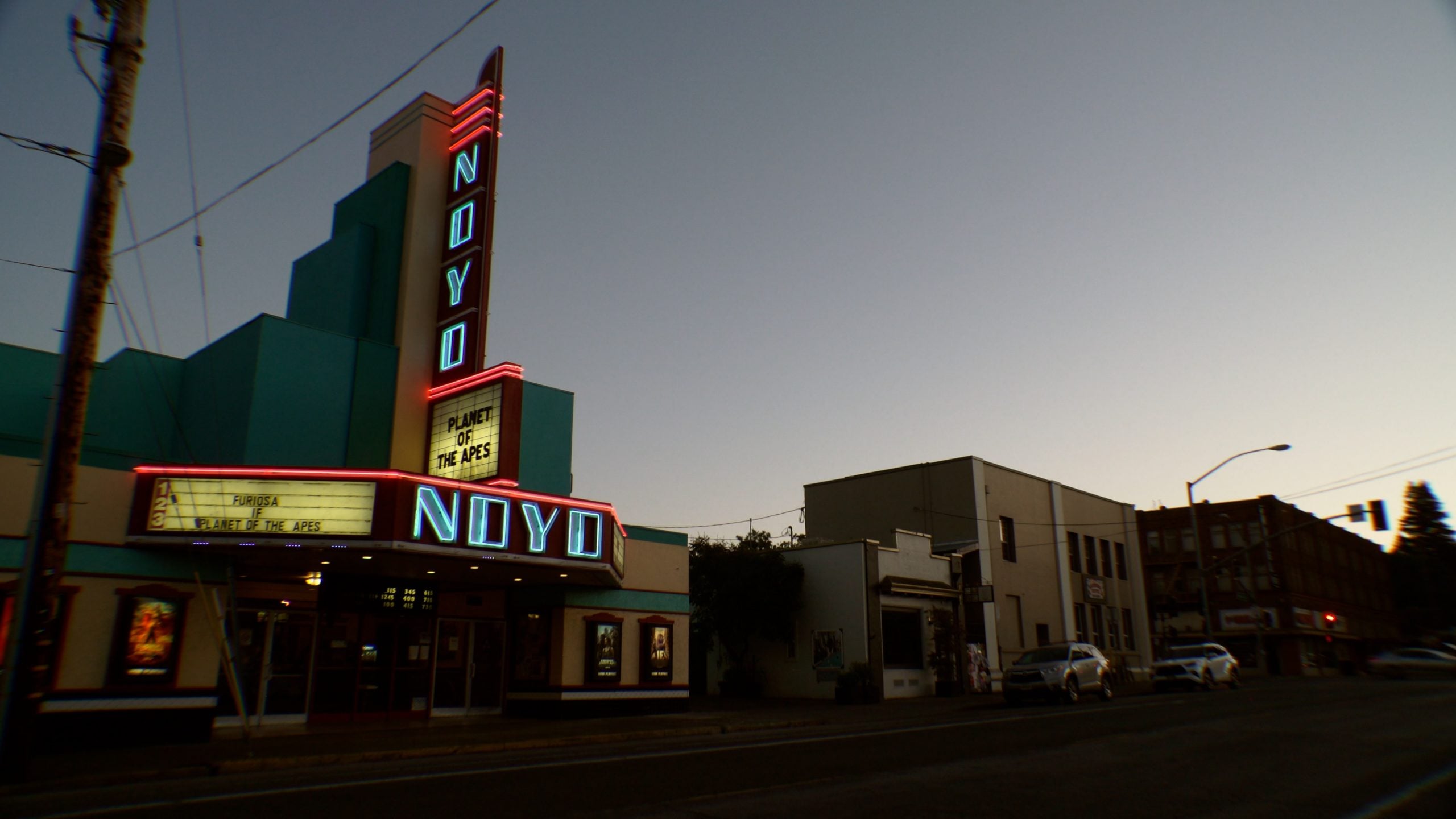 Street view of the neon-lit Noyo Theater at dusk with "Planet of the Apes" on the marquee, surrounded by nearby buildings and parked cars.