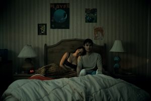 A young woman and man sit on a bed in a dimly lit bedroom, with the woman resting her head on the man's shoulder; posters and lamps are visible on the wall and nightstands.