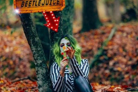 How Fantasy Films Continue To Inspire Real-World Fashion Person with green hair and striped shirt holds a candy bar in a forest. A lit "Beetlejuice" sign with a red arrow points down at them. Autumn leaves cover the ground.