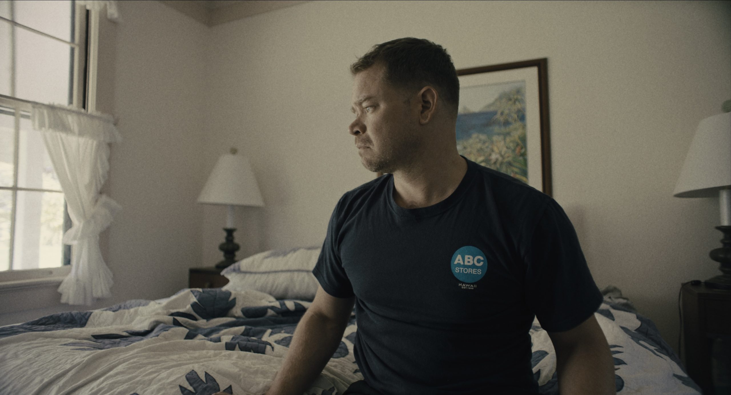 Man in an "ABC Stores" t-shirt sits on a bed in a sunlit bedroom, looking out a window.