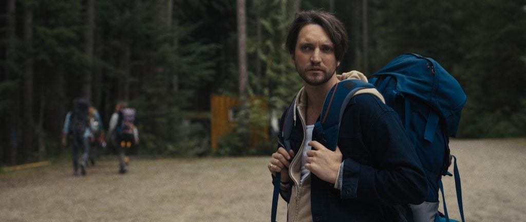 A man with a blue backpack stands outdoors in a wooded area, looking to the side, while three hikers walk away in the background.