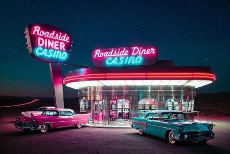 A retro diner and casino lit by neon signs at night, with two vintage cars parked outside under a starry sky.