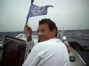 Person in a white shirt smiles at the camera while holding a blue flag on a small boat in the ocean under a cloudy sky.