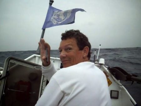 Person in a white shirt smiles at the camera while holding a blue flag on a small boat in the ocean under a cloudy sky.