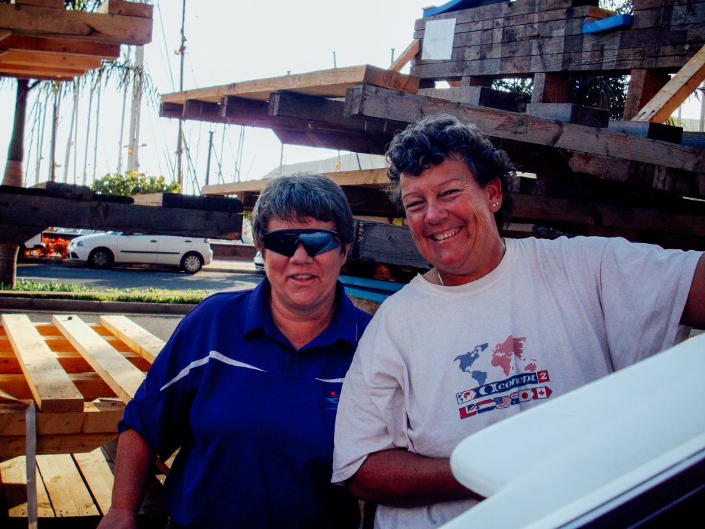 Two people stand outdoors by stacked wooden pallets, smiling at the camera. One wears sunglasses and a polo shirt; the other wears a printed T-shirt. Daylight and cars are visible in the background.