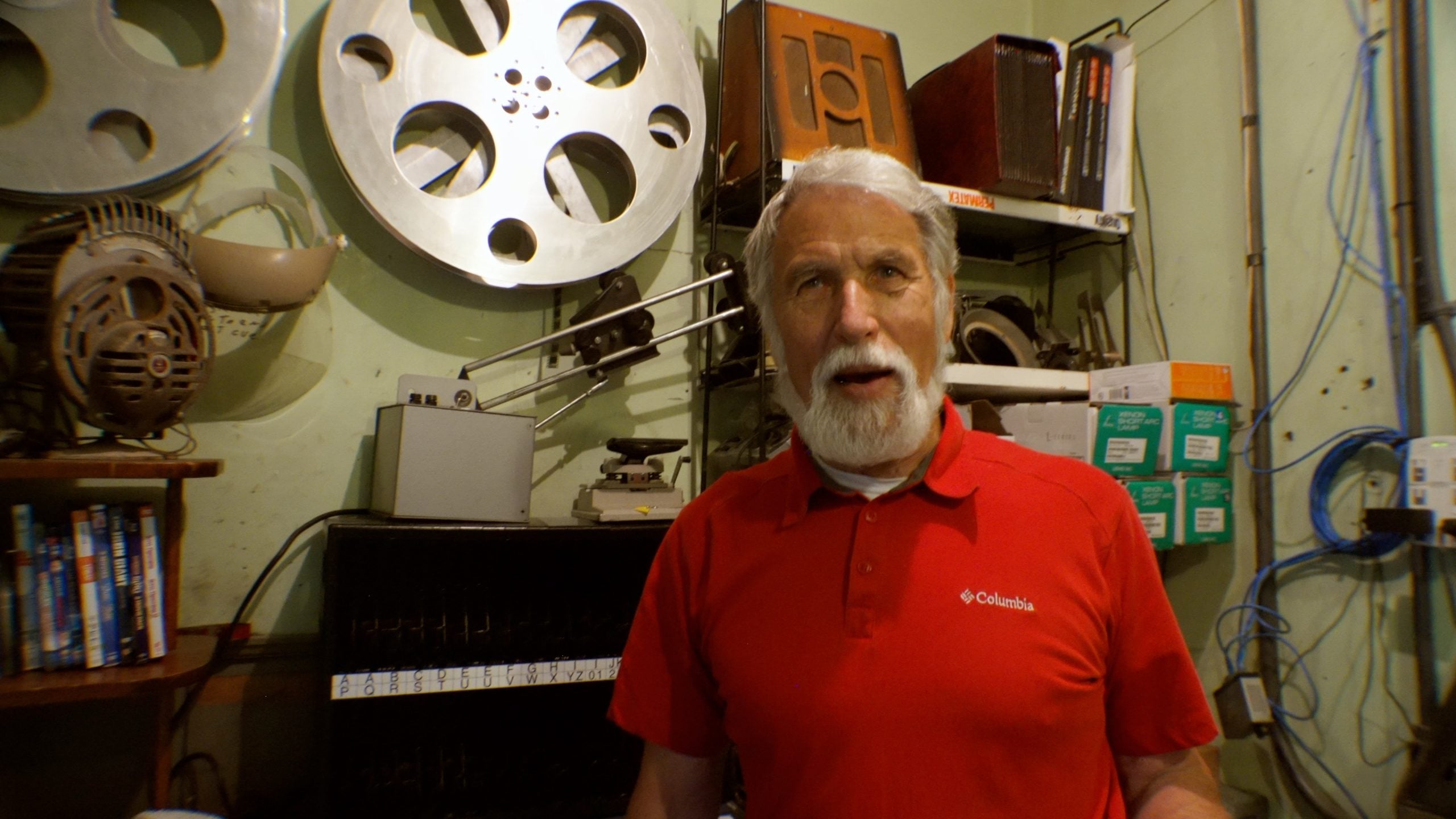 An older man with a white beard and red polo shirt stands in a room with film reels, electronics, and shelves of books and boxes.
