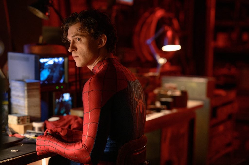 A person in a Spider-Man suit sits at a cluttered desk in a dimly lit room, looking over their shoulder. Computer screens and various objects are visible in the background.