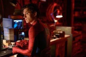 A person in a Spider-Man suit sits at a cluttered desk in a dimly lit room, looking over their shoulder. Computer screens and various objects are visible in the background.