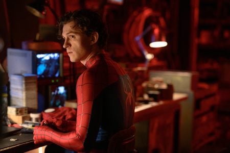 A person in a Spider-Man suit sits at a cluttered desk in a dimly lit room, looking over their shoulder. Computer screens and various objects are visible in the background.
