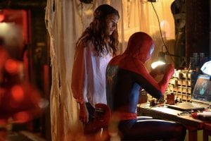 A woman stands next to Spider-Man, who is seated at a cluttered workbench repairing his suit, with tools and a laptop in a dimly lit room.