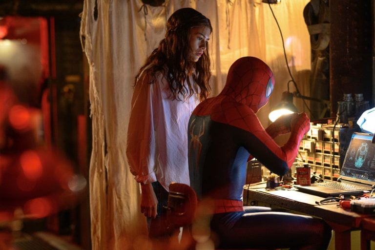 A woman stands next to Spider-Man, who is seated at a cluttered workbench repairing his suit, with tools and a laptop in a dimly lit room.